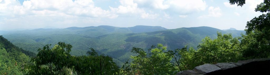 A wide view of forested mountain ridges under summer clouds, seen from a stone overlook