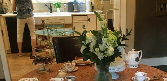 A dining table set for English tea with floral china and white flowers, the hostess at work in the kitchen beyond