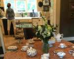 A dining table set for English tea with floral china and white flowers, the hostess at work in the kitchen beyond